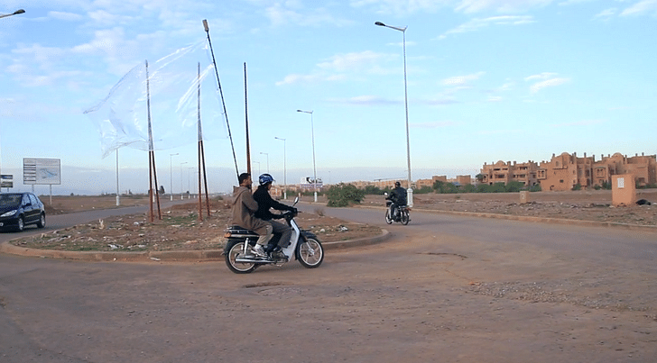In 'Place of Dead Roads,' the two riders aimlessly loop around an unfinished racetrack, an abandoned project by the Moroccan government. The transparent flag seems to take on the architecture it covers, implicitly demanding an enframent that simultaneously escapes grasp. Credit: Melia/Bitelli