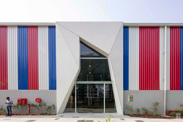 The massive rectilinear enclosure has walls that are patterned in bright red, blue & white bands with a central parametric-inspired main entrance doorway into the space. 
