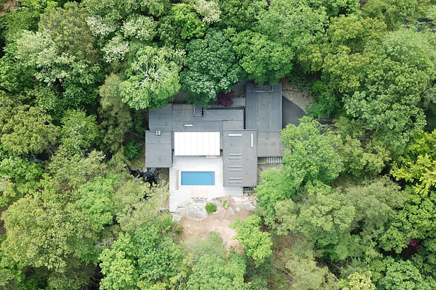 An aerial drone photo shows the house nestled in the wood landscape and the small stream that runs under the living room.