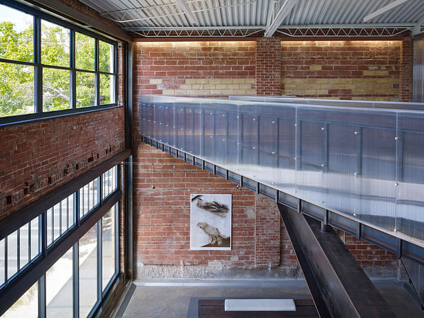 The entry lobby overlook showing historic beam pockets on the left, the 1930s bowstring truss roof, and the new steel and polycarbonate bridge.